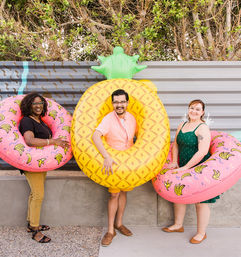 Three people posing on an urban patio with colorful inflatable pool floats — a giant pineapple float and two pink banana-pattern donut floats — in front of a corrugated metal fence and leafy shrubs.