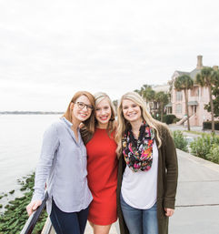 Three smiling friends posing on a waterfront promenade by palm trees and pastel historic homes — one in a red dress, two in casual tops and jeans, bright overcast sky