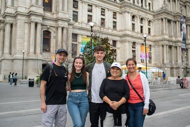 Five smiling adults posing on a downtown plaza in front of a grand historic city hall building with ornate stone columns and colorful banners