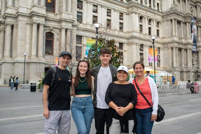 Five smiling adults posing on a downtown plaza in front of a grand historic city hall building with ornate stone columns and colorful banners