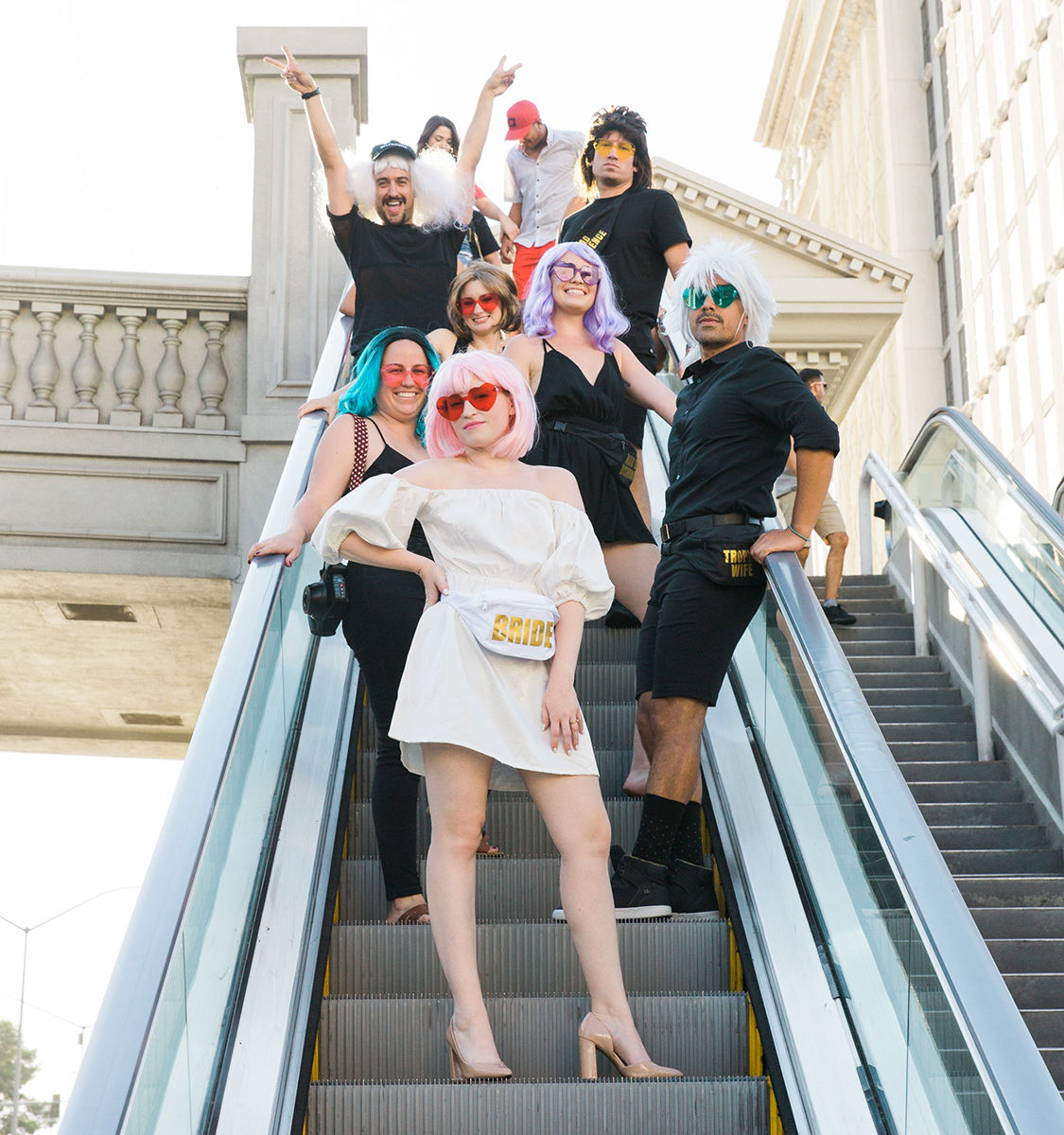 Bachelorette party group in colorful wigs and sunglasses posing on an outdoor city escalator, bride in a white dress and pink wig wearing a 'BRIDE' fanny pack leading the playful crew.