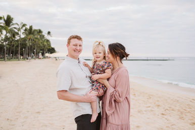 Happy parents holding a toddler on a palm-lined sandy beach by the calm ocean and pier under a soft cloudy sky