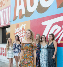 Six women laughing in summer dresses posed against a colorful urban mural reading "DON'T MESS WITH", with a woman in a floral off-shoulder dress front and center