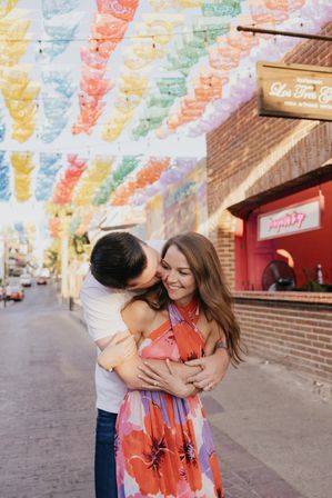 Couple embracing and smiling on a vibrant pedestrian street under colorful papel picado banners, woman in bright floral dress beside brick storefronts.