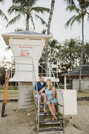 Couple sitting barefoot on steps of a white lifeguard tower at a tropical beach with palm trees and sand; tower sign reads NO LIFEGUARD ON DUTY CALL 911.