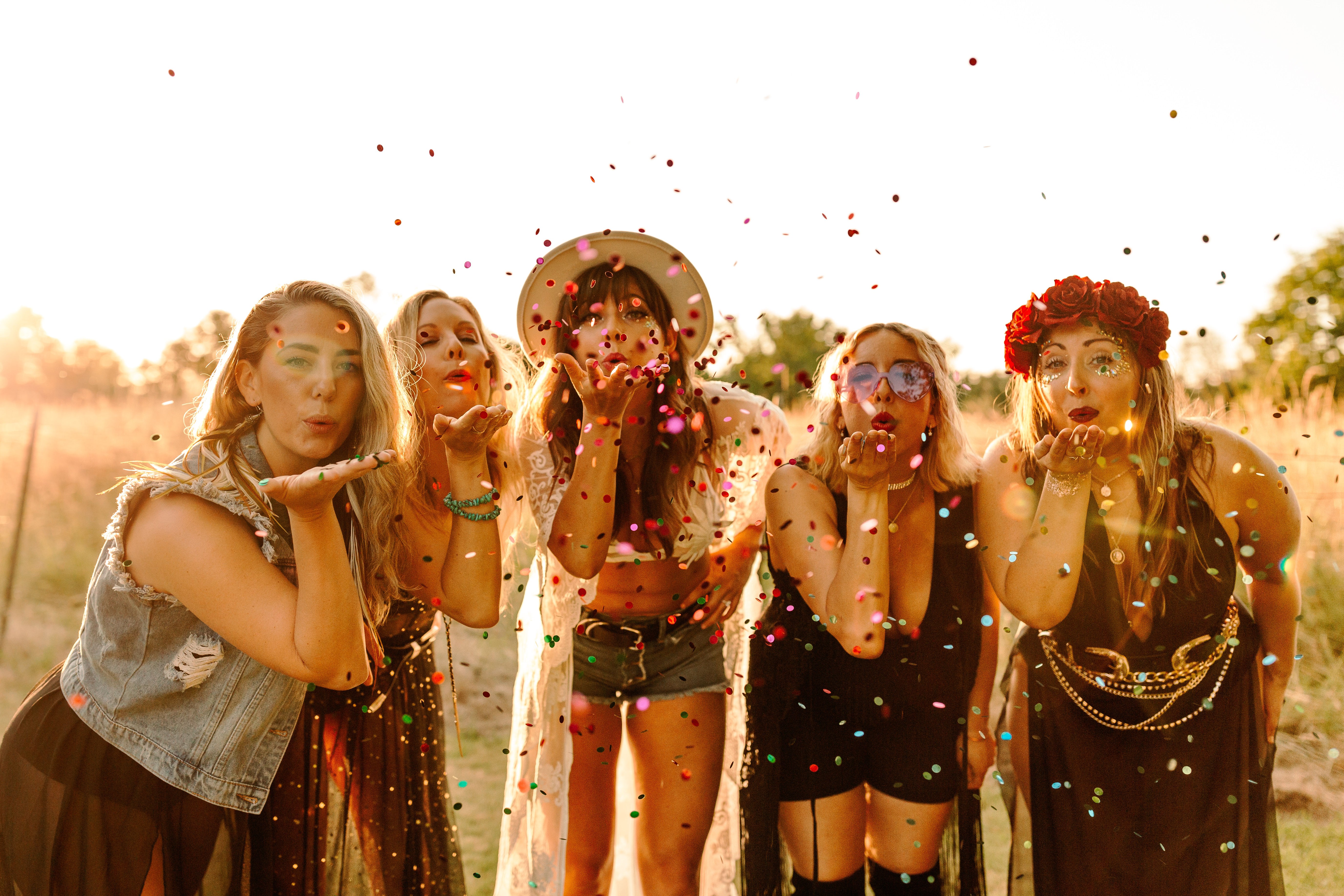 Five women in boho festival outfits blowing colorful confetti toward the camera in a sunlit grassy field at sunset, playful outdoor celebration.