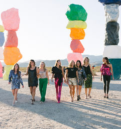 Group of eight women in colorful party outfits walking past towering multicolored stacked-rock sculptures in a sunny desert art installation