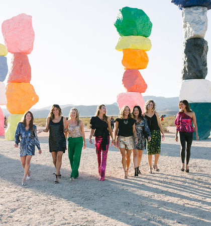 Group of eight women in colorful party outfits walking past towering multicolored stacked-rock sculptures in a sunny desert art installation