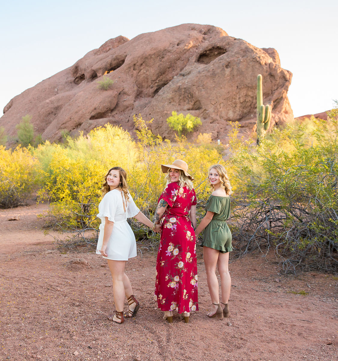 Three women in summer dresses holding hands and looking back in a sunlit desert portrait with yellow scrub, a saguaro cactus, and a large red rock formation