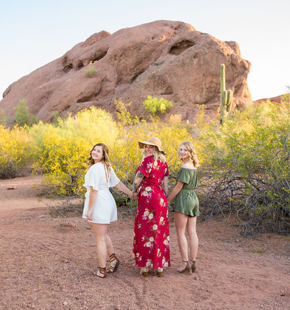 Three women in summer dresses holding hands and looking back in a sunlit desert portrait with yellow scrub, a saguaro cactus, and a large red rock formation