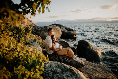 Couple embracing on a rocky shoreline at golden hour, sitting among boulders and looking out over rippling water toward distant mountains.