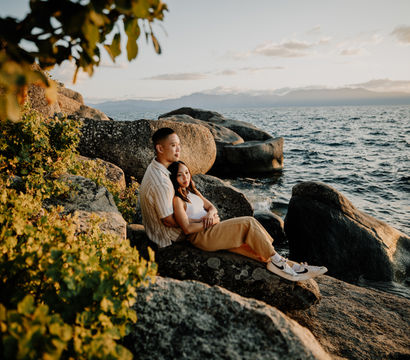Couple embracing on a rocky shoreline at golden hour, sitting among boulders and looking out over rippling water toward distant mountains.