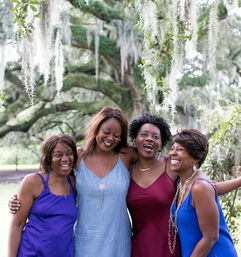 Four joyful friends in colorful sundresses laughing and hugging beneath a Spanish-moss-draped live oak in a sunlit park