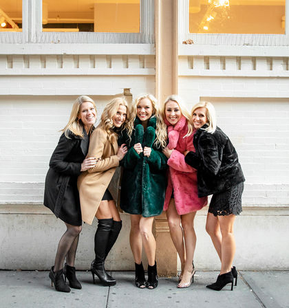 Five smiling women in winter coats (camel, black, green, pink) huddled arm‑in‑arm on a city sidewalk in front of a white brick building, wearing heels in a playful urban group portrait.