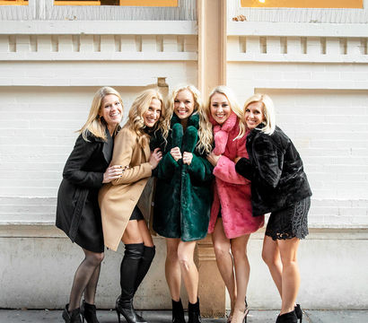Five smiling women in colorful faux-fur coats and heels posing on a downtown sidewalk in front of a white brick storefront