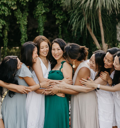 Seven women in summer dresses smiling and sharing a joyful group hug around a friend in a green dress in a lush outdoor garden.