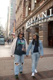 Two people holding hands and smiling as they stroll down a busy downtown sidewalk past an ornate stone theater entrance, both in denim, ripped jeans and sunglasses.