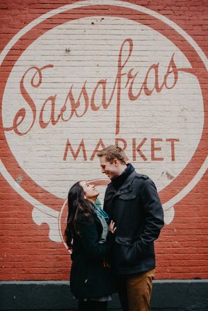 Smiling couple in winter coats share a laugh in front of a large circular brick-wall mural featuring a stylized market sign — urban street photo.