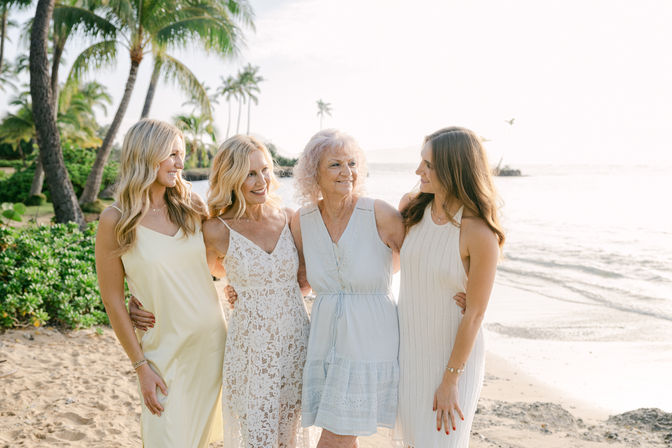 Four smiling women across generations in light summer dresses embracing on a sunny tropical beach with palm trees and gentle ocean waves
