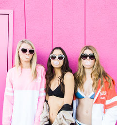 Playful summer trio in bikinis and heart-shaped sunglasses blowing bubblegum in front of a bright pink textured wall