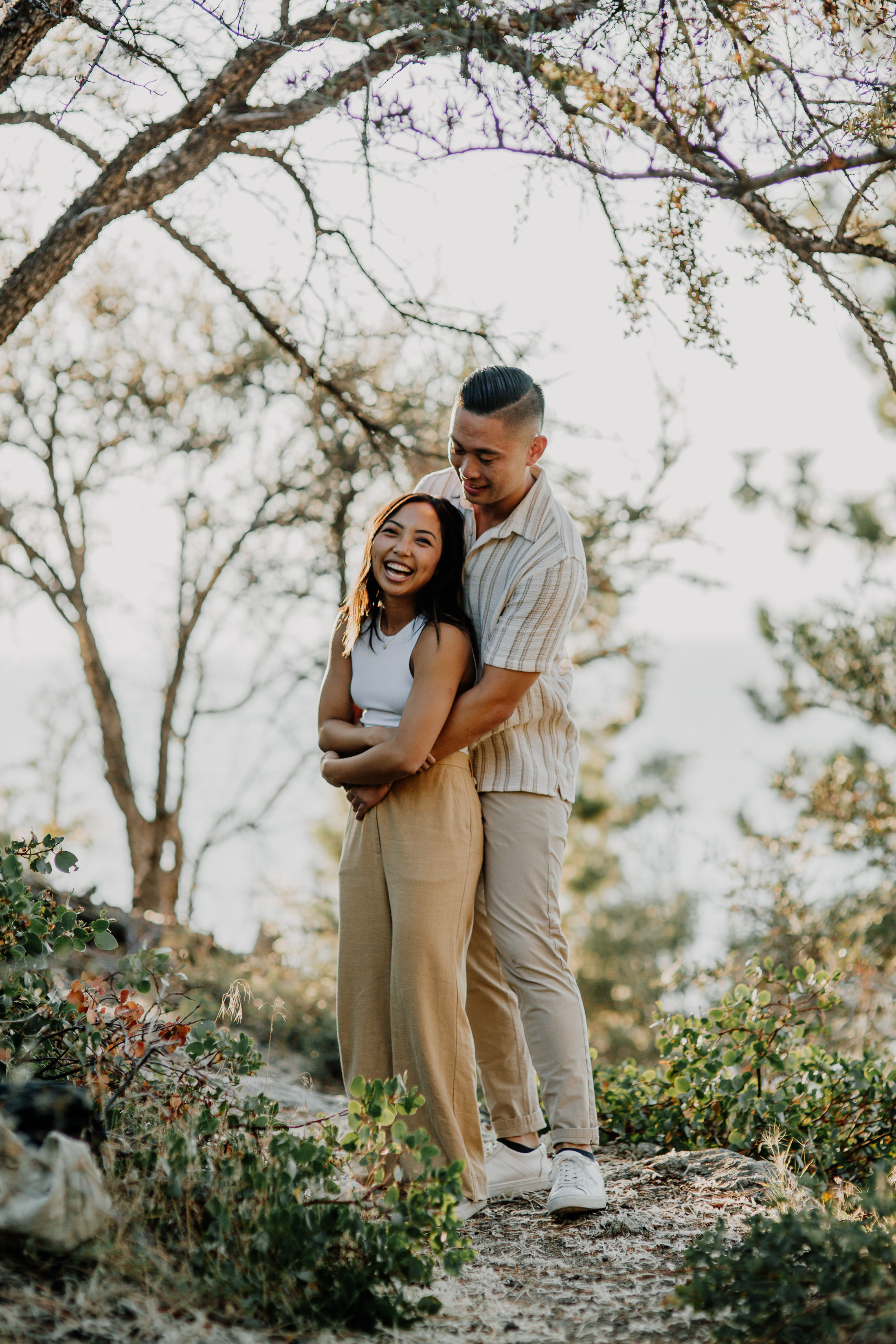 Playful young couple hugging on a sunlit forest trail, woman laughing as her partner embraces her from behind among trees and low shrubs