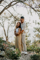 Playful young couple hugging on a sunlit forest trail, woman laughing as her partner embraces her from behind among trees and low shrubs