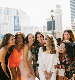 Seven laughing women in colorful summer outfits posing together on a sunny Las Vegas promenade with high-rise hotels and decorative street lamps in the background.