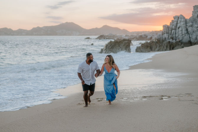 Smiling couple holding hands and running barefoot along a sandy beach at sunset, waves lapping the shore with rocky coastline and distant mountains in the background.