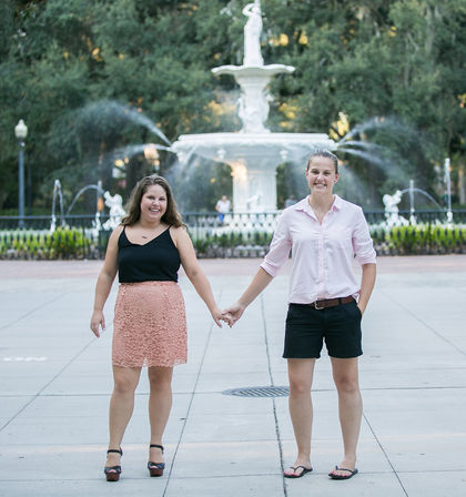 Two women holding hands and smiling in front of an ornate white fountain at a city park plaza, with water sprays and trees in the background.