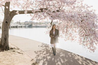 Person strolling beneath pink cherry blossom branches along a sunlit waterfront path in spring, wearing an oversized coat and knee-high boots with soft water reflections in the background.