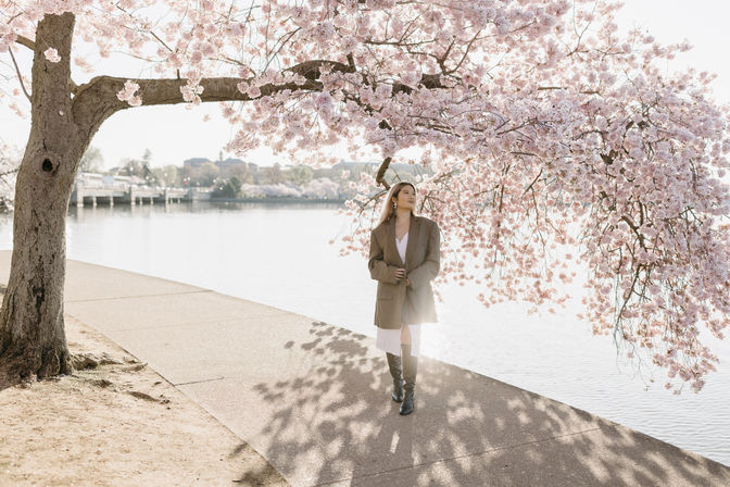 Person strolling beneath pink cherry blossom branches along a sunlit waterfront path in spring, wearing an oversized coat and knee-high boots with soft water reflections in the background.