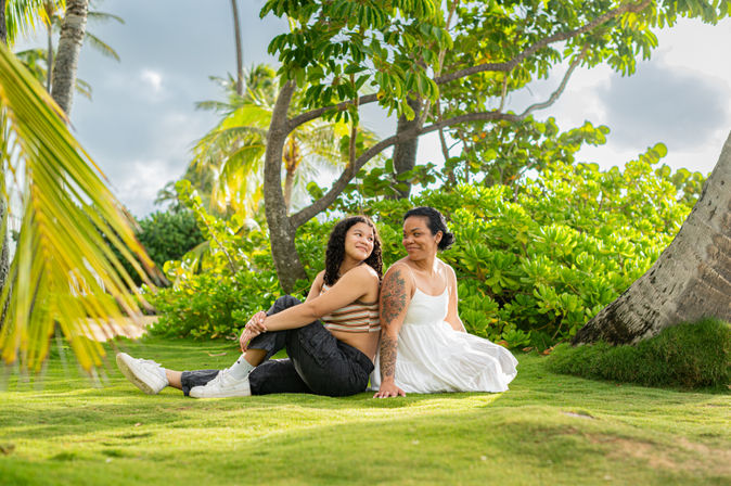 Two people sitting back-to-back on a sunlit grassy lawn under palm trees in a tropical setting, one in a white sundress with an arm tattoo and the other in a striped crop top, black pants and white sneakers, smiling amid lush green foliage.
