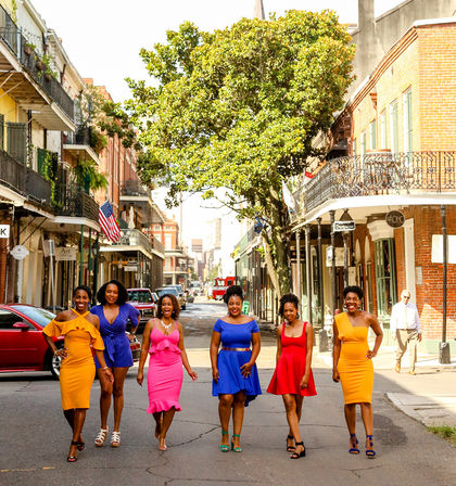 Six women in bright summer dresses walking confidently down a sunny New Orleans French Quarter street lined with brick buildings, wrought-iron balconies, and a large shade tree.