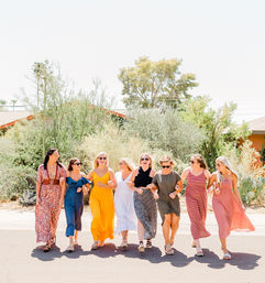 Eight women in colorful summer dresses walking arm-in-arm, smiling and laughing on a sunny suburban street lined with cacti and desert shrubs