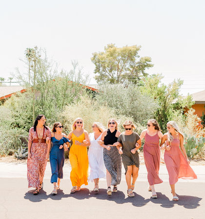 Eight women in colorful summer dresses walking arm-in-arm, smiling and laughing on a sunny suburban street lined with cacti and desert shrubs