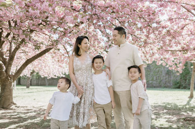 Smiling family of five — parents and three young boys — posing beneath blooming pink cherry blossom trees in a sunlit park for a spring family portrait.