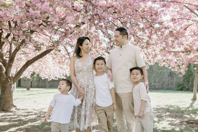 Smiling family of five — parents and three young boys — posing beneath blooming pink cherry blossom trees in a sunlit park for a spring family portrait.