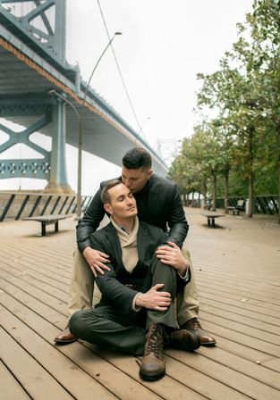 Romantic couple — two men in smart-casual outfits embrace on a riverside wooden boardwalk beneath a steel suspension bridge, tree-lined urban waterfront on an overcast day.