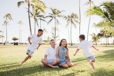 Happy family of four at a sunny palm-lined beachfront park — parents seated on the grass laughing while two boys run and play barefoot
