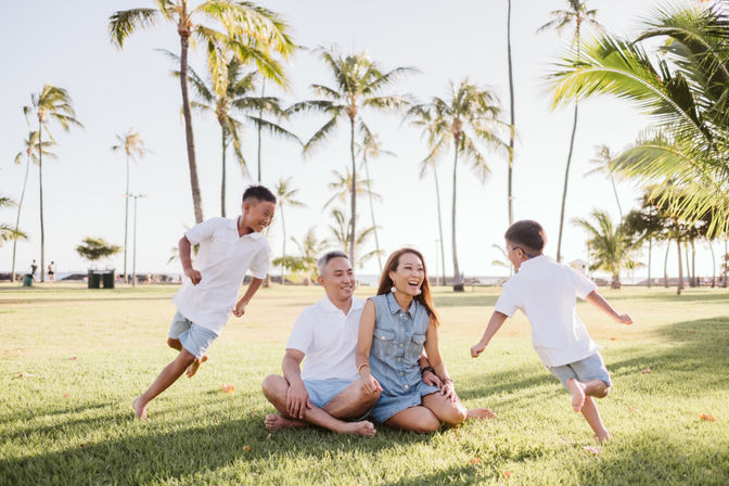 Happy family of four at a sunny palm-lined beachfront park — parents seated on the grass laughing while two boys run and play barefoot