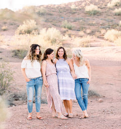 Four friends laughing together on a sunlit desert hillside with scrub brush, wearing casual summer dresses, jeans and sandals — outdoor group portrait