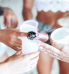 Close-up of hands pouring sparkling rosé into white plastic cups at a sunny beach party, friends toasting.