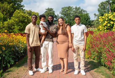 Smiling family of five on a sunny brick garden path lined with vibrant yellow and red flowers; father holds a toddler while mother and two teen sons stand beside.
