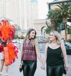 Two friends holding hands and smiling as they walk the Las Vegas Strip at dusk, with a red-feathered showgirl and hotel towers in the background.