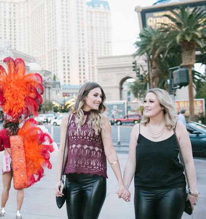 Two friends holding hands and smiling as they walk the Las Vegas Strip at dusk, with a red-feathered showgirl and hotel towers in the background.