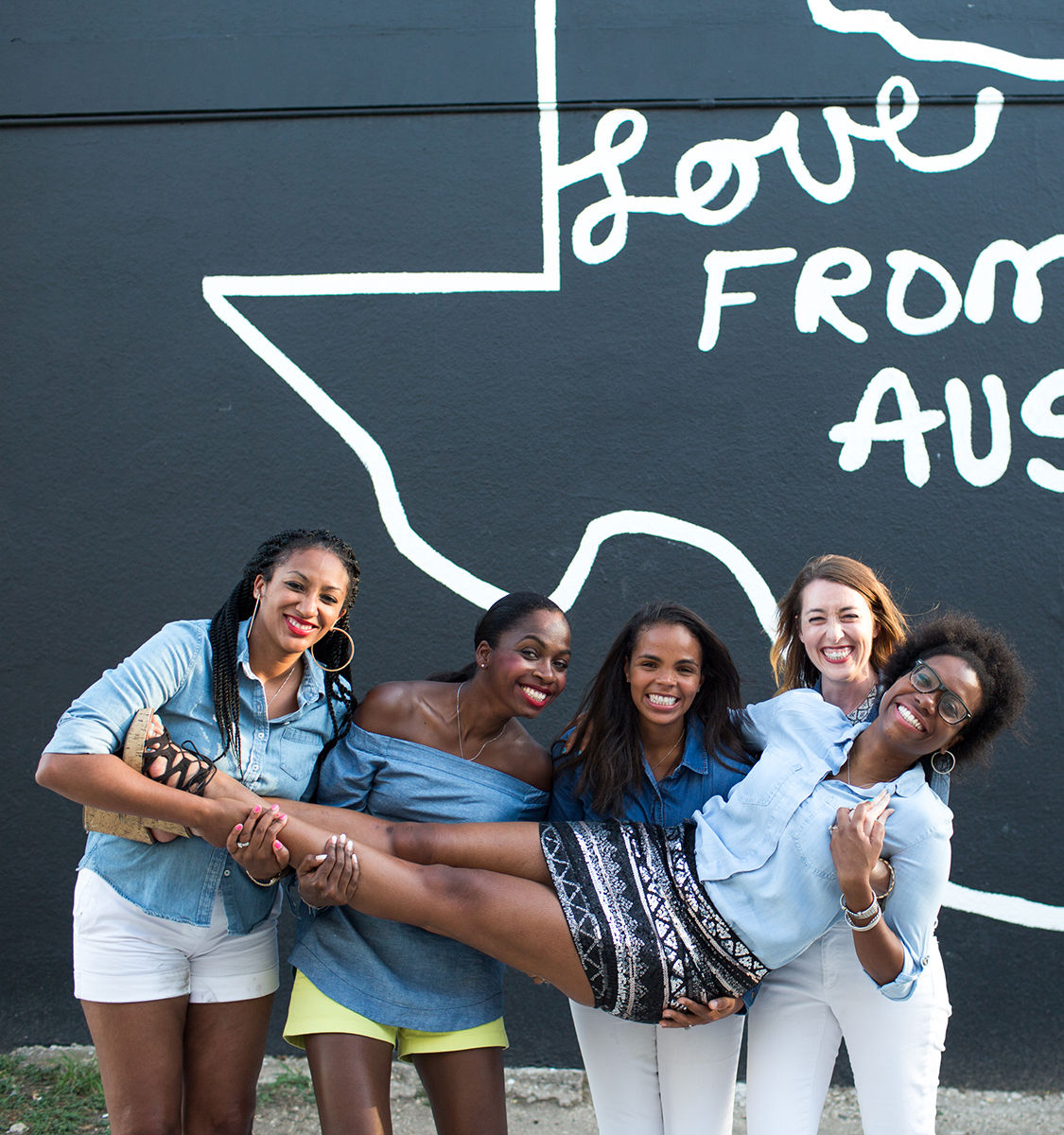 Five smiling women hold a friend horizontally in front of a black 