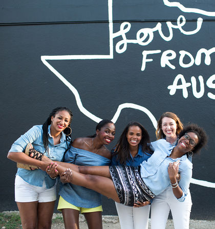 Five smiling women hold a friend horizontally in front of a black 