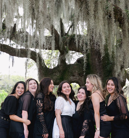 Seven women laughing together in black and white outfits beneath a sprawling southern live oak draped with Spanish moss — outdoor group portrait in a lush park setting.