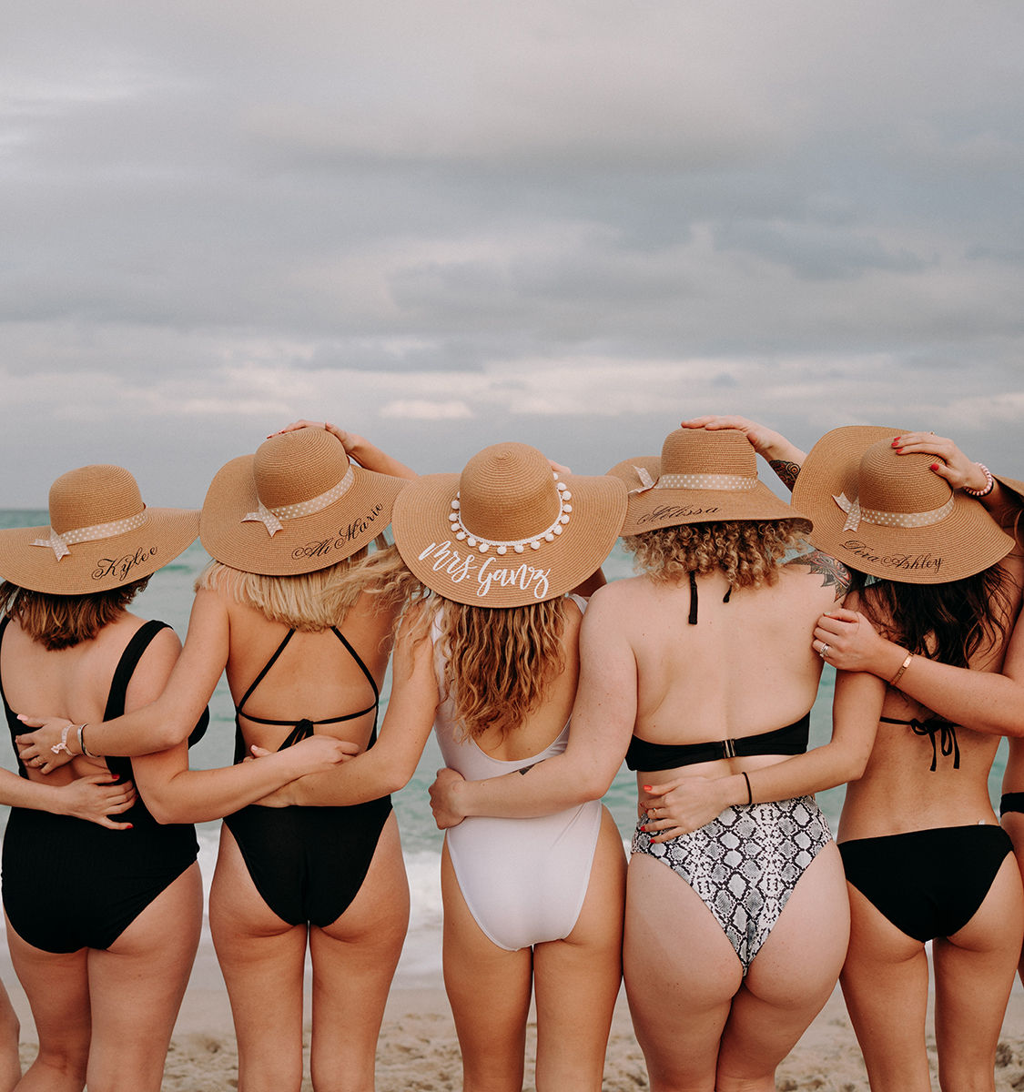 Six friends in swimsuits standing arm‑in‑arm on a sandy beach facing the ocean, wearing matching wide‑brimmed straw sun hats under a cloudy sky.
