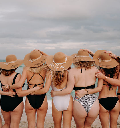 Six friends in swimsuits standing arm‑in‑arm on a sandy beach facing the ocean, wearing matching wide‑brimmed straw sun hats under a cloudy sky.